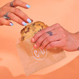 Choc Chip New York Cookie held by hands with rings in a Crumbs & Doilies bag against peach background
